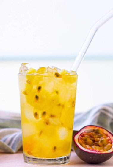 a glass of Pineapple Refresher with ice and a straw next to a half of a passion fruit
