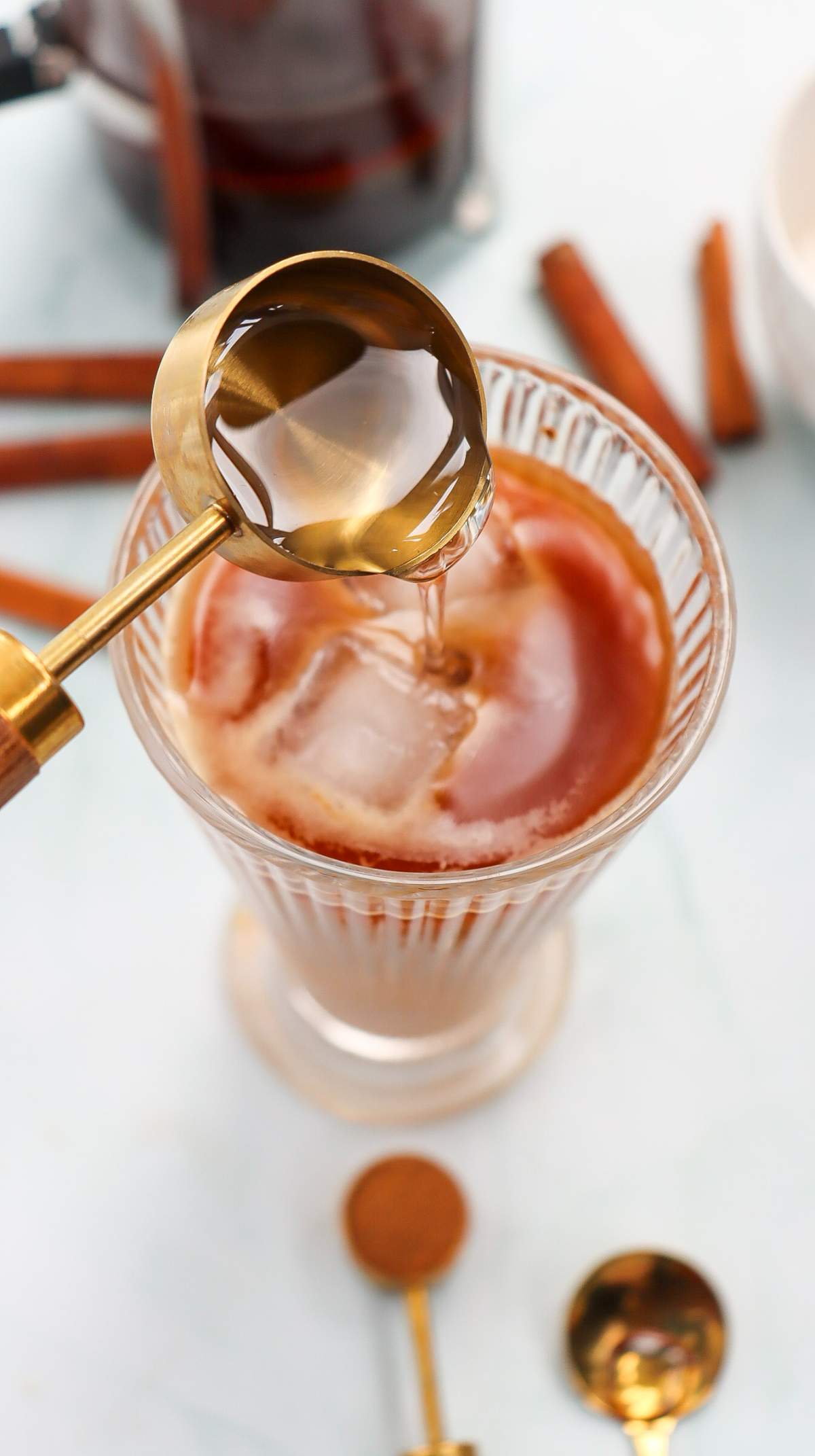 close-up, top-down shot of a Starbucks-inspired Iced Cinnamon Dolce Latte in a ribbed glass filled with ice, as golden syrup is being poured from a gold measuring spoon.