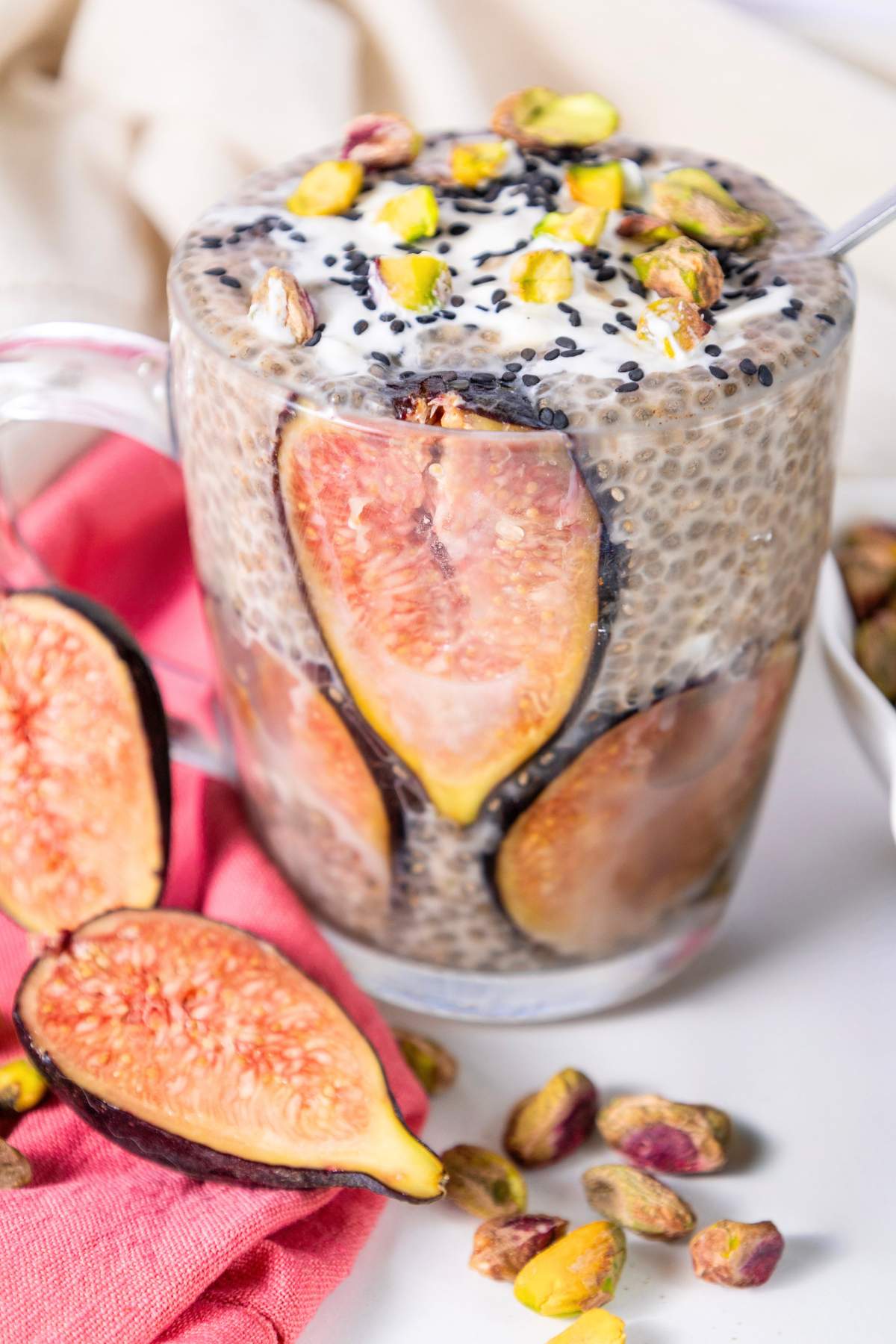 lose-up of a fig chia pudding served in a clear glass, with fresh figs lining the sides of the glass. The pudding is topped with yogurt, pistachios, and black sesame seeds.