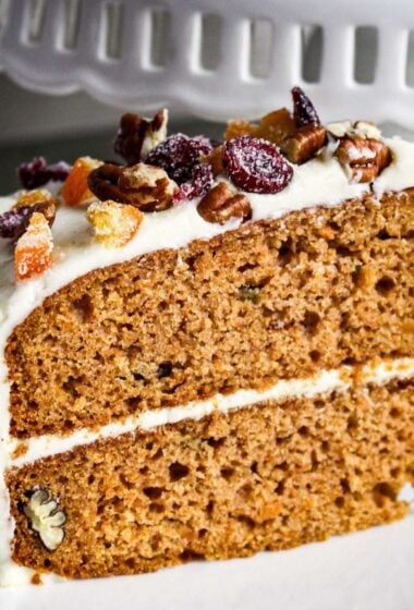 close-up of a slice of moist sweet potato cake with cream cheese frosting, topped with chopped pecans and dried cranberries.