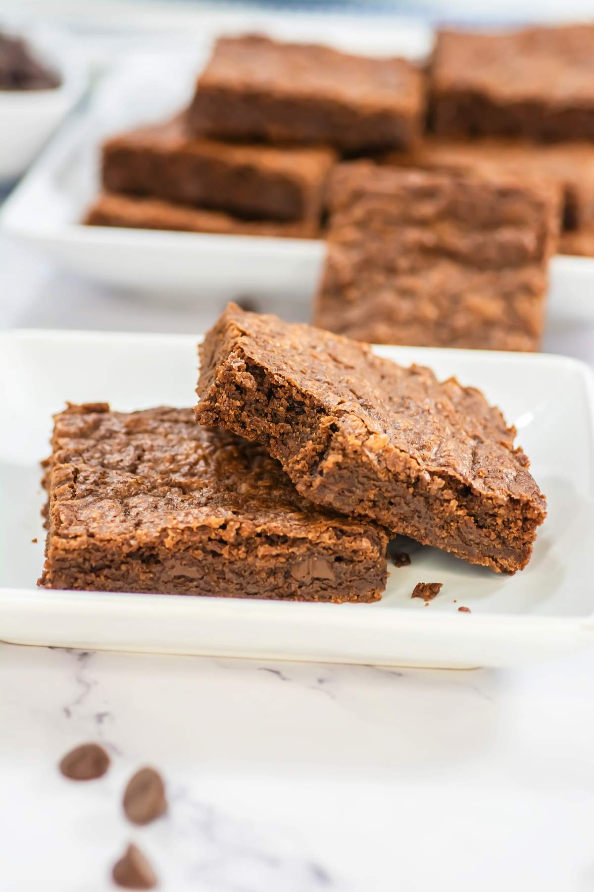 Two Sheet Pan Brownies stacked on a white plate, one resting at an angle to reveal the soft, fudgy center and melted chocolate chips, with more brownies blurred in the background and scattered chips on a marble surface.