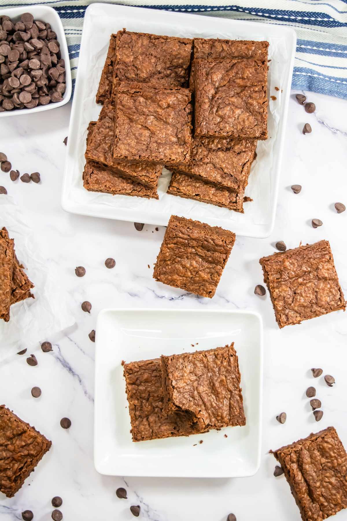 Overhead view of sliced Sheet Pan Brownies on white plates, surrounded by scattered chocolate chips on a marble surface, with a bowl of chocolate chips and a striped kitchen towel in the background.