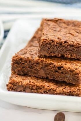 Close-up of three stacked Sheet Pan Brownies on a white plate lined with parchment paper, showing their rich, dense texture and gooey chocolate chips, with a bowl of chocolate chips blurred in the background.