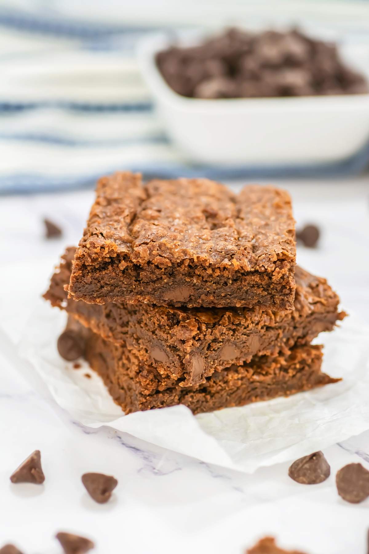 Stack of chewy Sheet Pan Brownies on white parchment paper, showing a rich, fudgy interior with melty chocolate chips, surrounded by scattered chocolate chips and a soft-focus striped towel and bowl in the background.