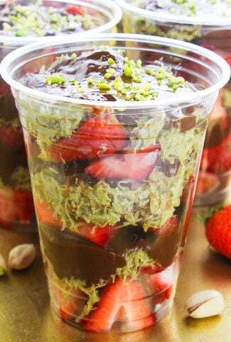 Plastic cups of Dubai Chocolate Strawberries filled with layers of sliced strawberries, shredded pistachio konafa, and rich chocolate ganache, topped with crushed pistachios, served on a golden tray.