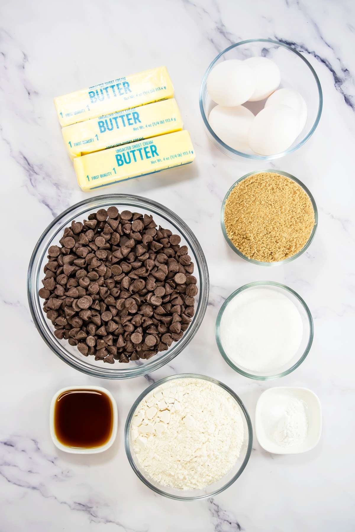 Overhead view of Sheet Pan Brownies ingredients on a marble surface, including sticks of butter, eggs, brown sugar, granulated sugar, chocolate chips, vanilla extract, flour, and baking powder.
