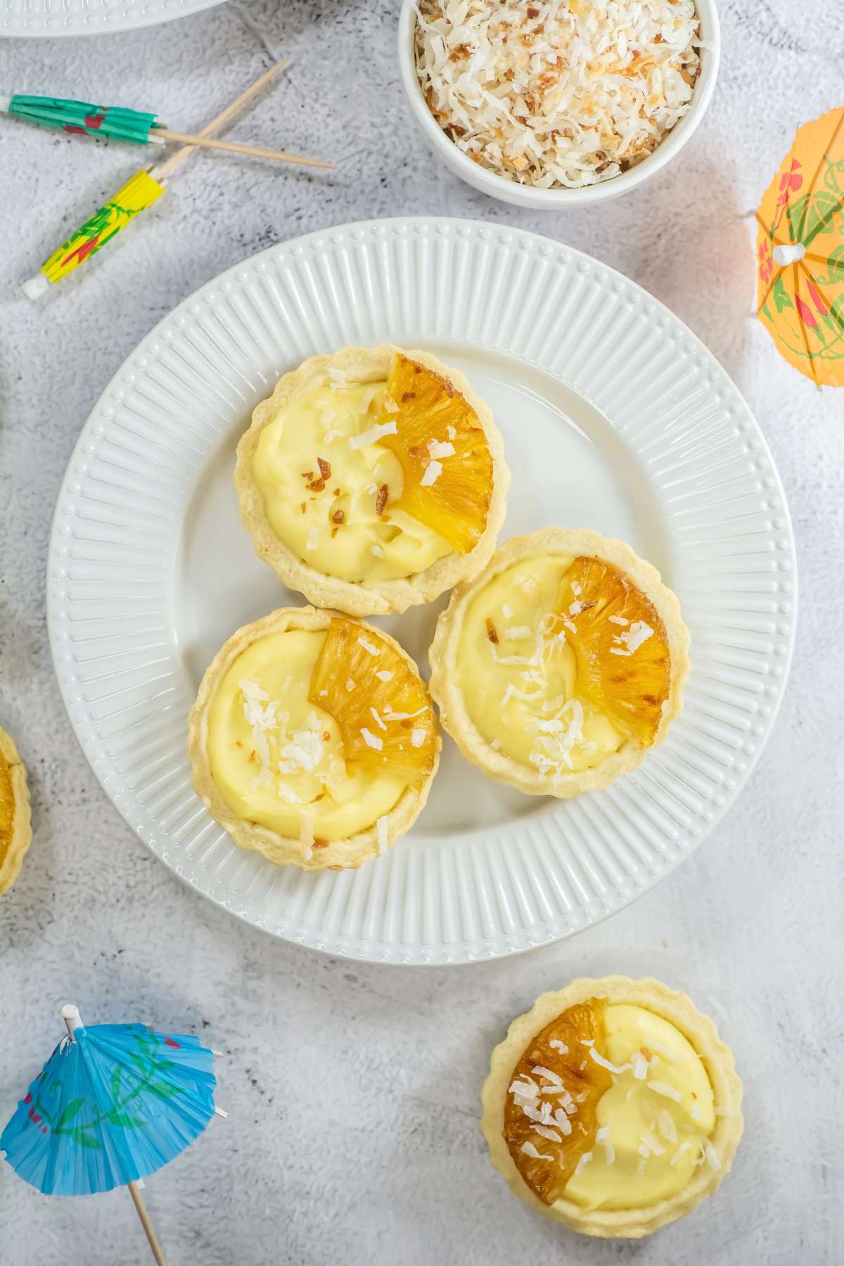 Top-down view of four mini piña colada tarts on a white plate, garnished with caramelized pineapple and shredded coconut, surrounded by tropical-themed props like cocktail umbrellas and a bowl of toasted coconut.