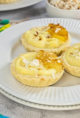 Close-up of three Piña Colada mini tarts on a white plate, filled with creamy coconut pineapple filling, topped with caramelized pineapple slices and shredded coconut, with a bowl of toasted coconut in the background.