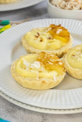 Close-up of three Piña Colada mini tarts on a white plate, filled with creamy coconut pineapple filling, topped with caramelized pineapple slices and shredded coconut, with a bowl of toasted coconut in the background.