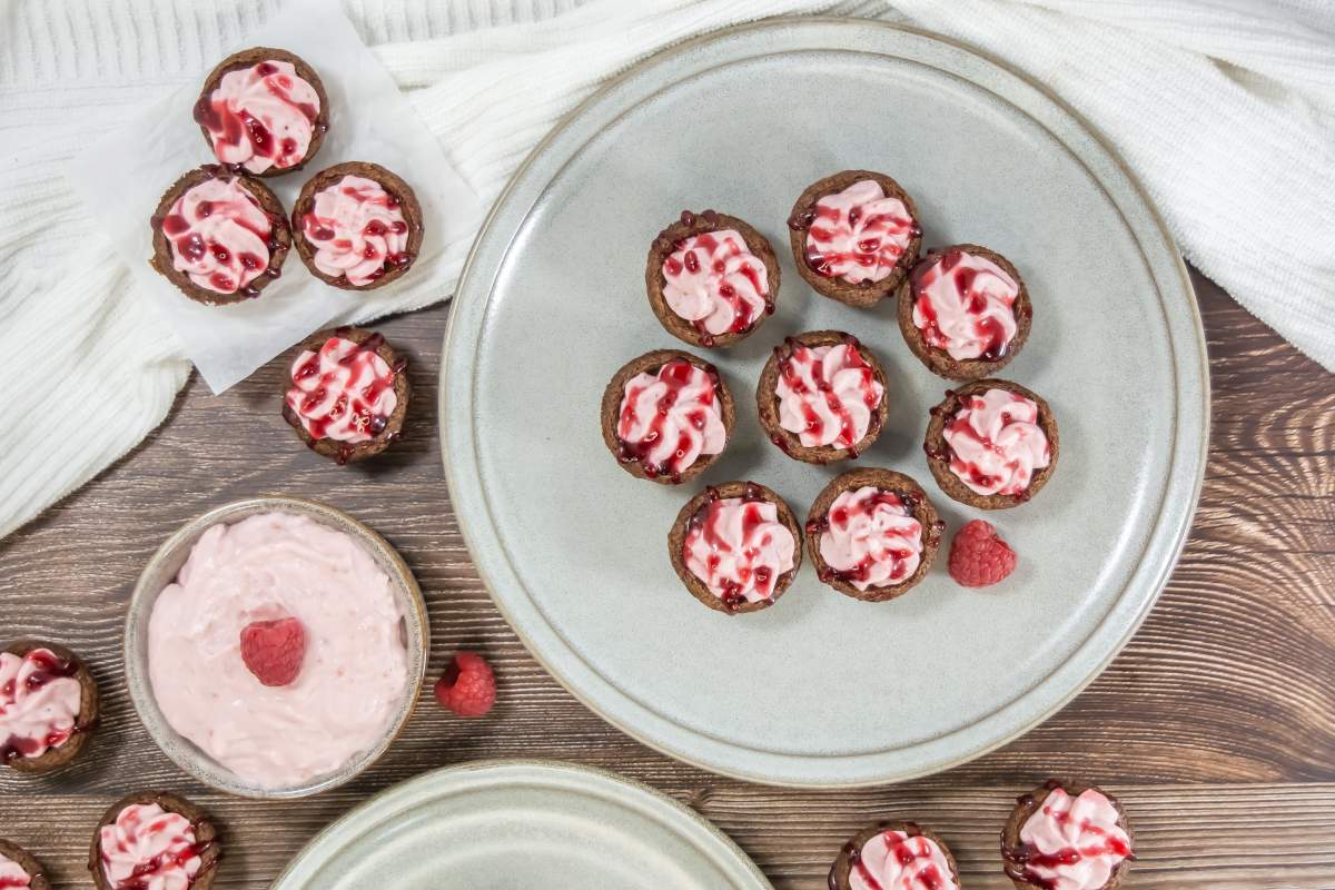 Overhead view of raspberry mousse brownie bites topped with pink raspberry mousse and raspberry sauce, arranged on a neutral ceramic plate with fresh raspberries, a bowl of mousse, and a white kitchen towel on a wooden surface.