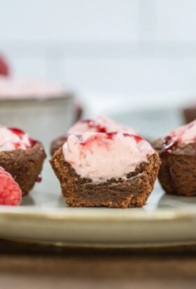 Close-up side view of a halved raspberry mousse brownie bite, showing layers of fudgy brownie and creamy pink mousse topped with raspberry sauce, with whole raspberries and more bites in the background.