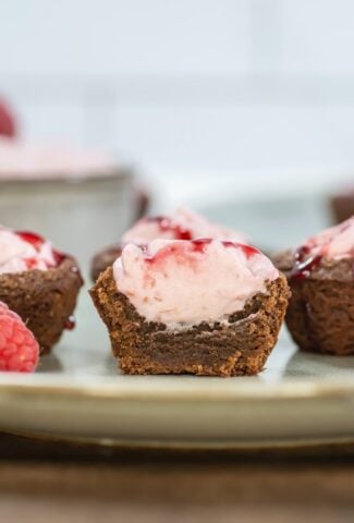 Close-up side view of a halved raspberry mousse brownie bite, showing layers of fudgy brownie and creamy pink mousse topped with raspberry sauce, with whole raspberries and more bites in the background.