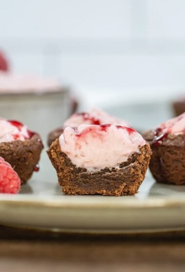 Close-up side view of a halved raspberry mousse brownie bite, showing layers of fudgy brownie and creamy pink mousse topped with raspberry sauce, with whole raspberries and more bites in the background.