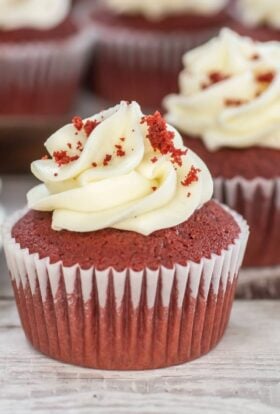 Close-up of a single red velvet cupcake topped with a generous swirl of cream cheese frosting and red cake crumbs, with more frosted cupcakes softly blurred in the background on a wooden surface.