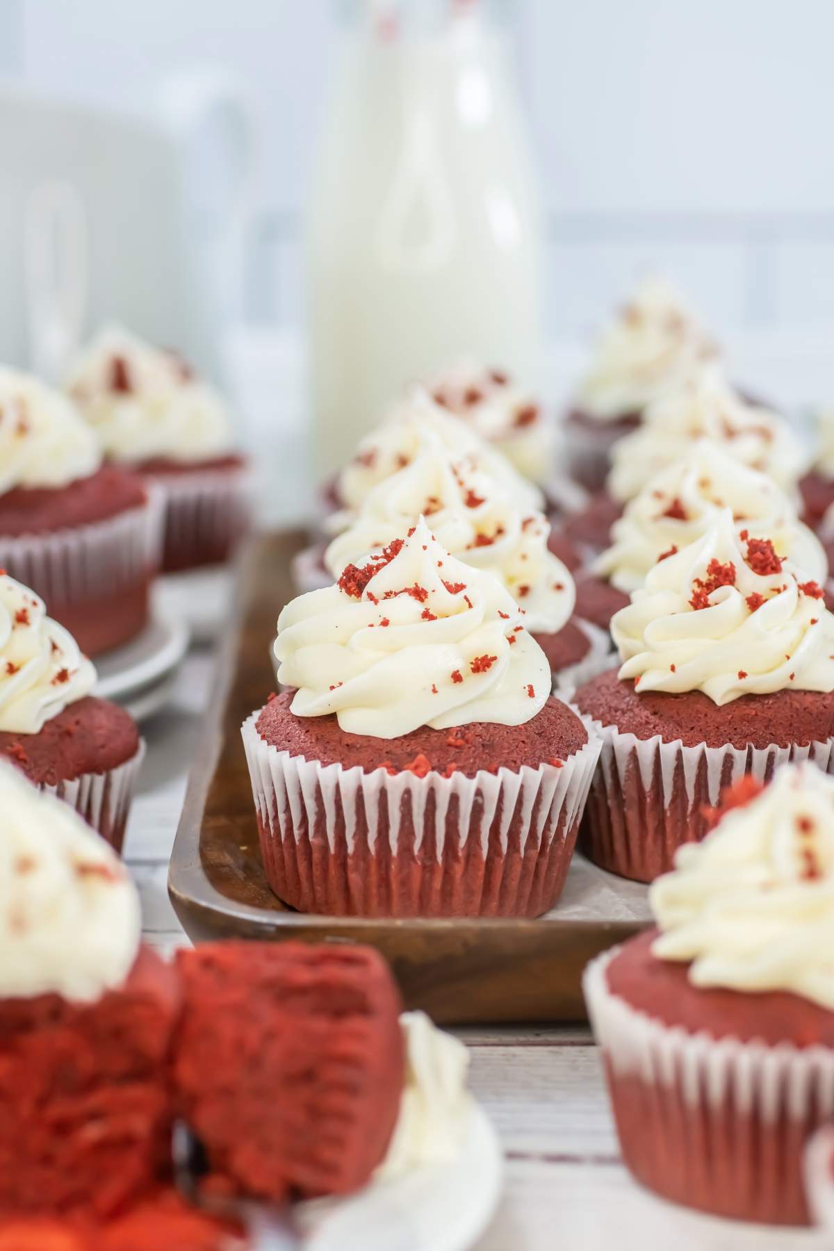 Close-up of red velvet cupcakes topped with swirls of cream cheese frosting and red cake crumbs, arranged on a wooden tray with a bottle of milk and more cupcakes in the background.
