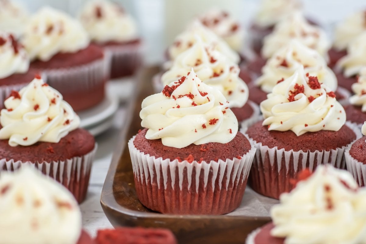 Tray of red velvet cupcakes topped with swirls of cream cheese frosting and red cake crumbs, with additional cupcakes in the background and foreground on a light wooden surface.