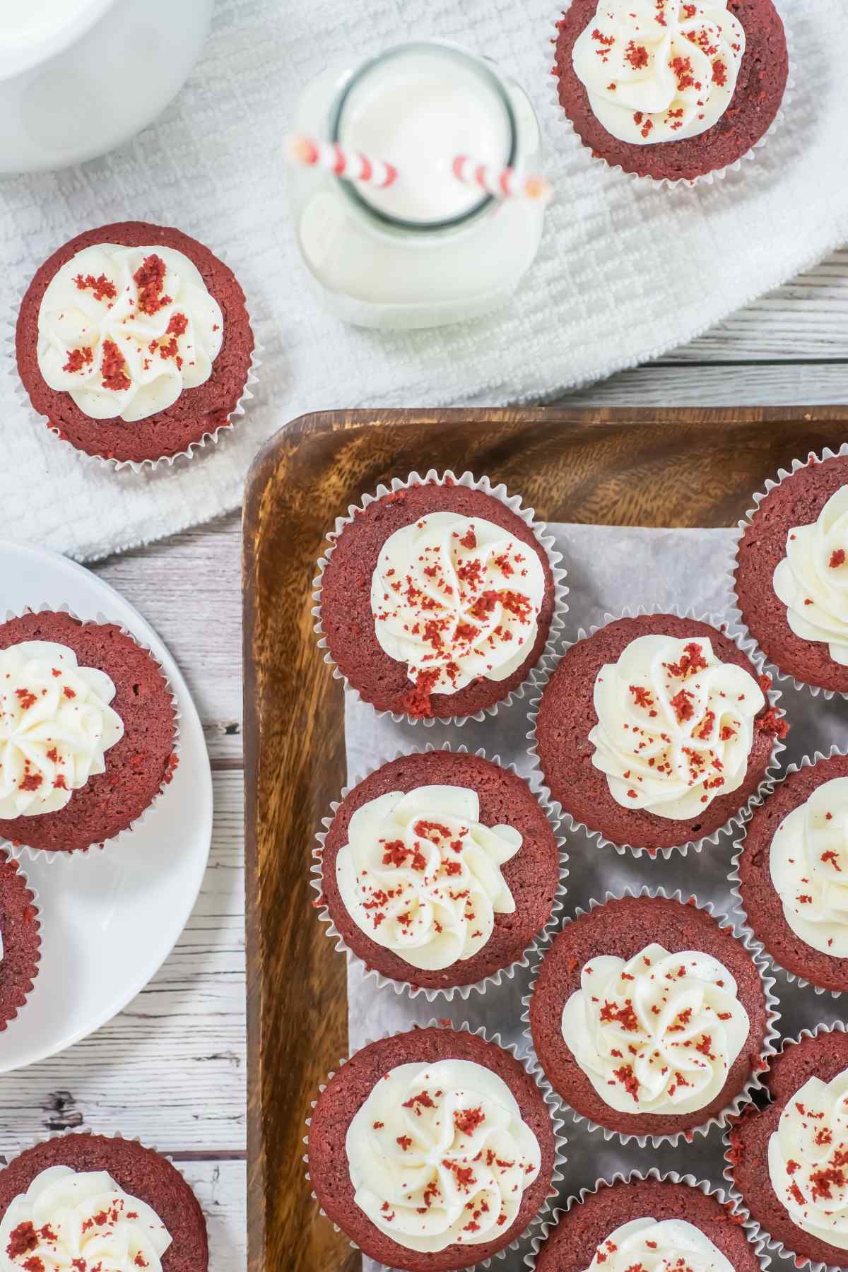Top-down view of red velvet cupcakes with cream cheese frosting and red cake crumbs, arranged on a wooden tray and white plate, with a bottle of milk and striped straws on a white towel.