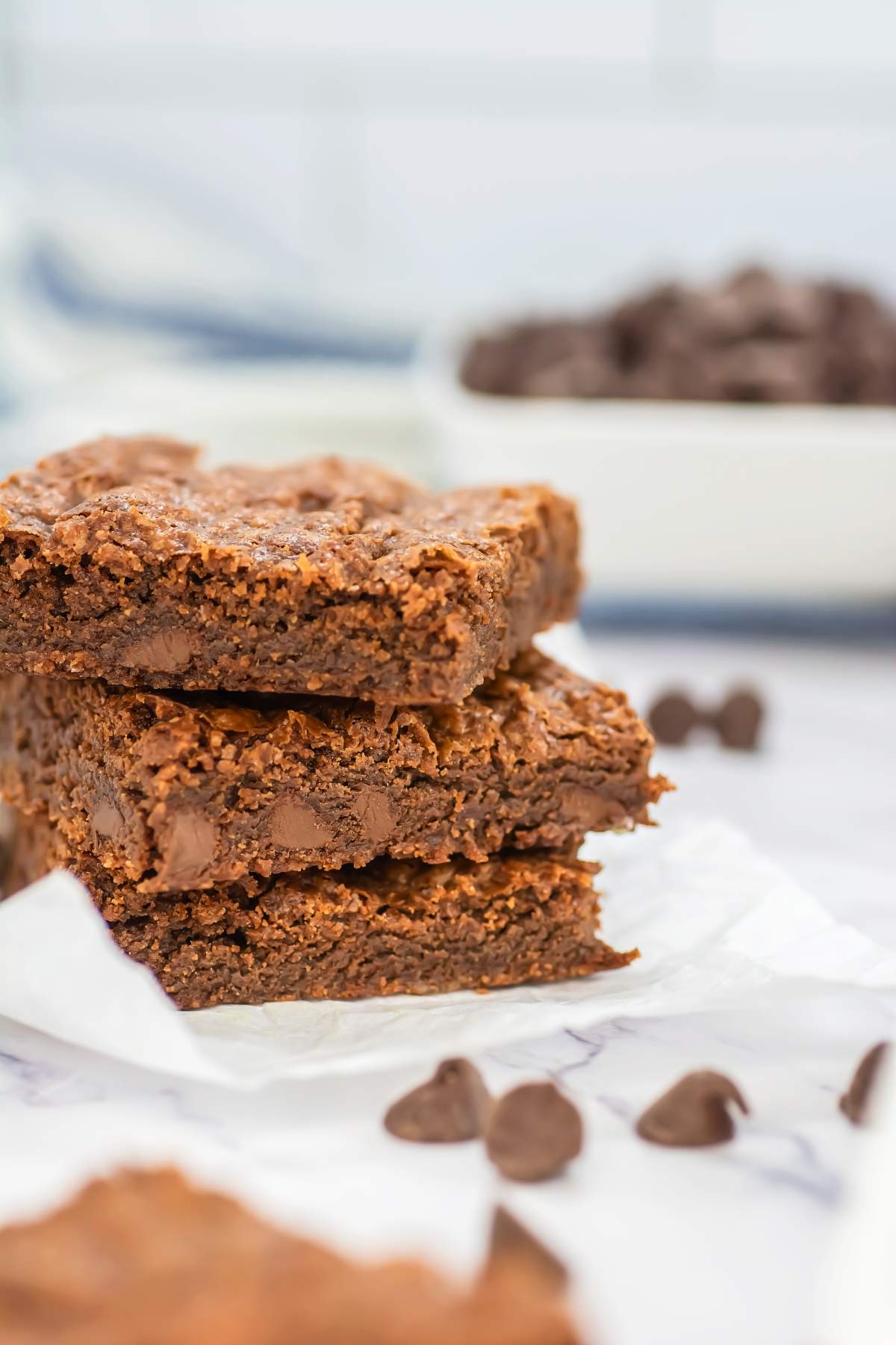 Close-up of a stack of gooey Sheet Pan Brownies on parchment paper, with a soft, fudgy texture and crisp edges, surrounded by scattered chocolate chips and a blurred bowl of chips in the background.